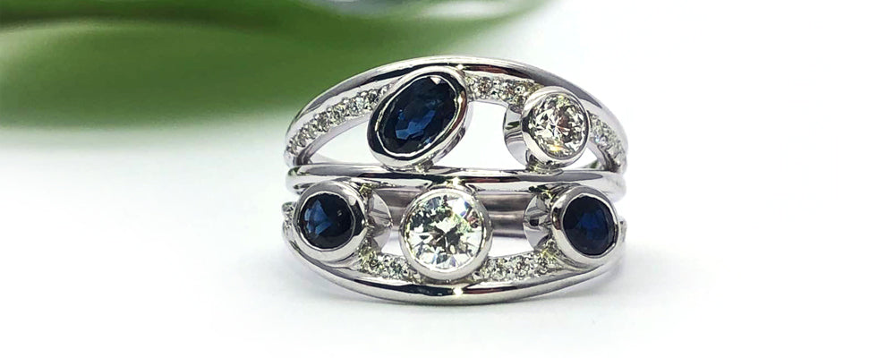 Bague sur mesure en or blanc au design ouvert et moderne, composée de plusieurs anneaux fusionnés en une seule pièce. Sertie de diamants de tailles variées et de saphirs bleus, la composition alterne sertis clos et pavés, offrant un jeu de volume et d’éclat.