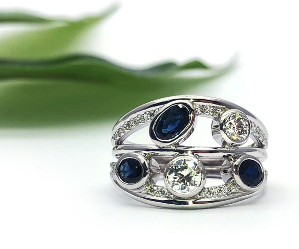 Bague sur mesure en or blanc au design ouvert et moderne, composée de plusieurs anneaux fusionnés en une seule pièce. Sertie de diamants de tailles variées et de saphirs bleus, la composition alterne sertis clos et pavés, offrant un jeu de volume et d’éclat.