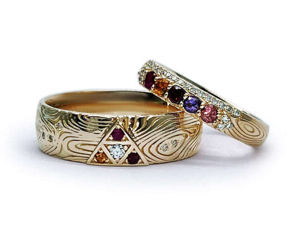 Ensemble de bagues en or jaune faites sur mesure. L’anneau homme, plus large, présente un motif triangulaire serti de diamant, grenat et rubis, sur un corps de bague texturé de style damas. L’anneau femme est pavé de pierres naturelles : grenat, spinelle, saphir violet, tourmaline rose et diamants.